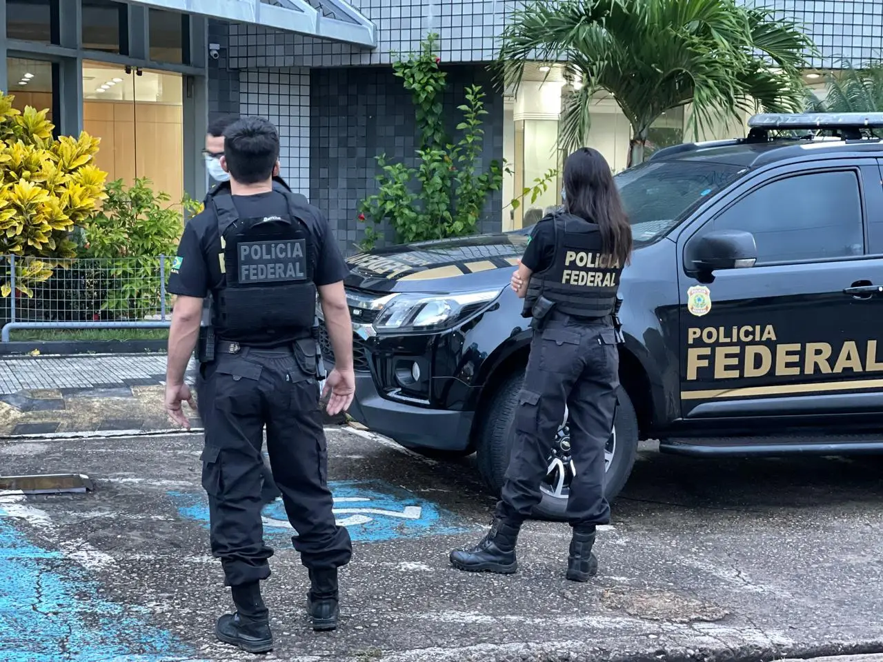 Agentes da Polícia Federal em ação ao lado de viatura oficial preta estacionada, com uniformes táticos e coletes à prova de balas.