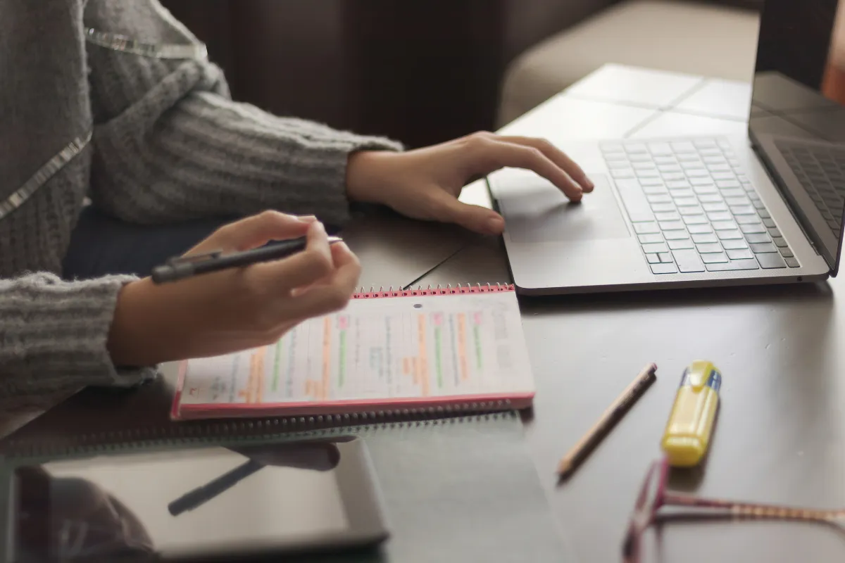 Pessoa estudando e escrevendo em caderno ao lado de um laptop, com caneta e outros materiais de estudo visíveis.
