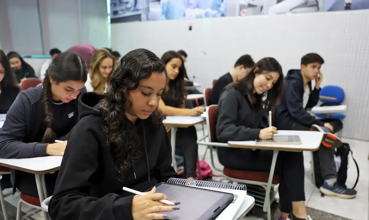 Estudantes concentrados usando tablets e cadernos em sala de aula moderna.