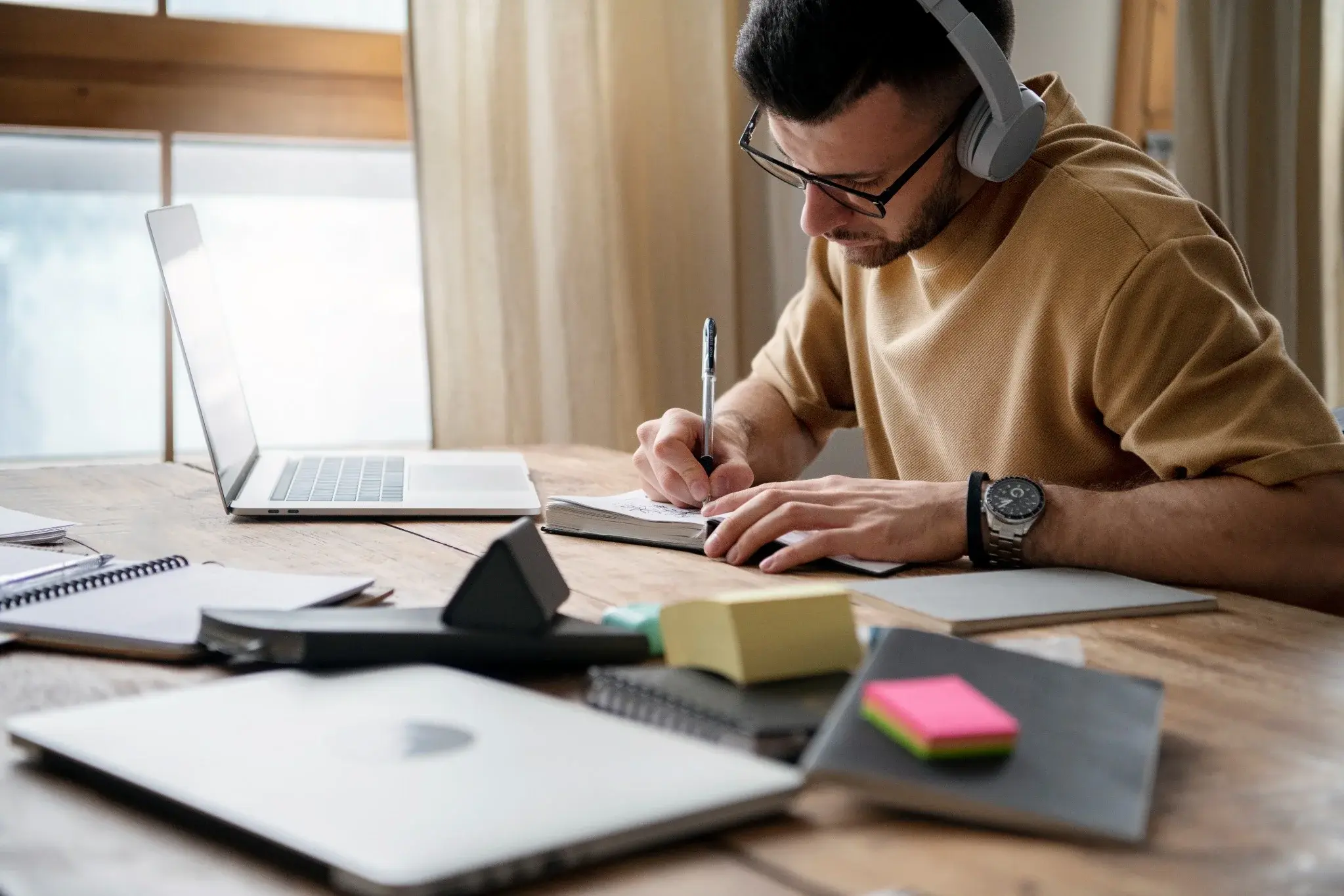 Jovem concentrado em preparação para concurso, estudando com laptop, fones de ouvido e materiais de apoio sobre a mesa.