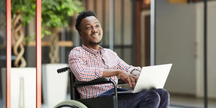 Homem em cadeira de rodas sorrindo e usando um laptop em um ambiente moderno.