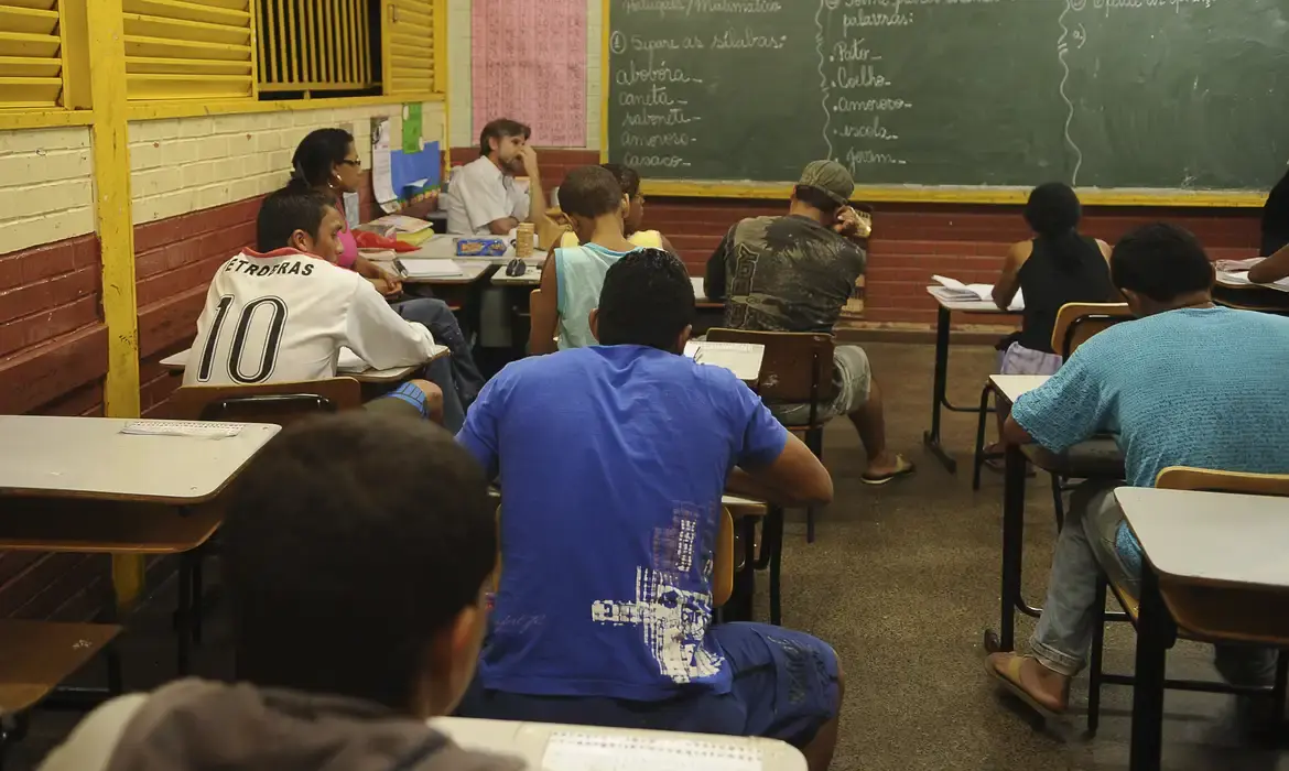 Alunos participando de aula em uma sala de ensino médio, com o professor ao fundo.