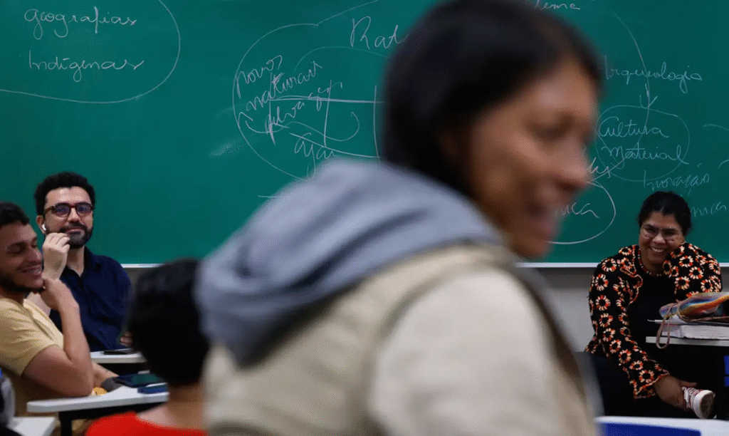 Alunos e professora em sala de aula com quadro verde ao fundo.