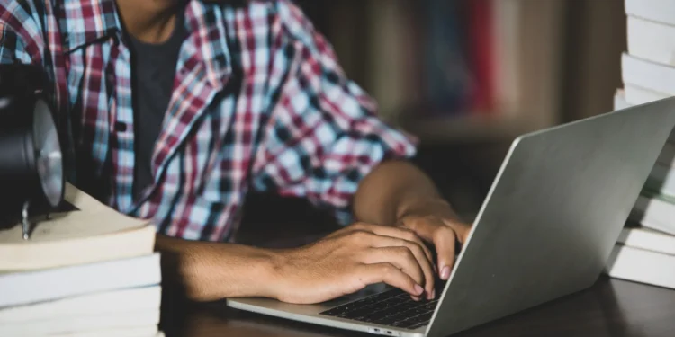 Jovem estudando com laptop na biblioteca cercado por livros.