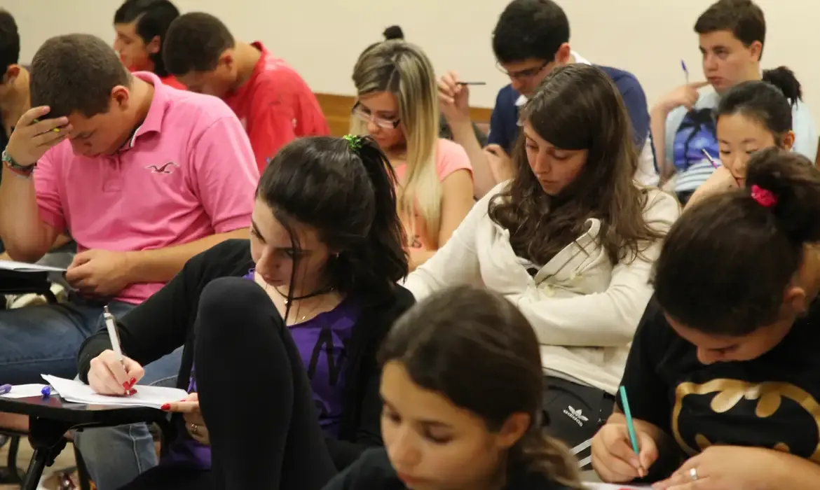 Estudantes concentrados realizando prova em sala de aula durante concurso.