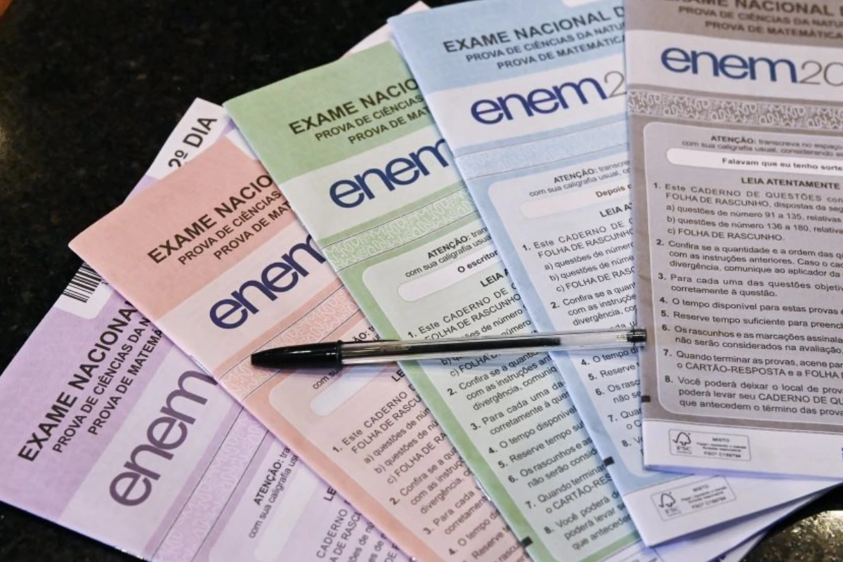 Provas do Exame Nacional do Ensino Médio organizadas em diferentes cores sobre uma superfície escura, com uma caneta preta sobre elas.