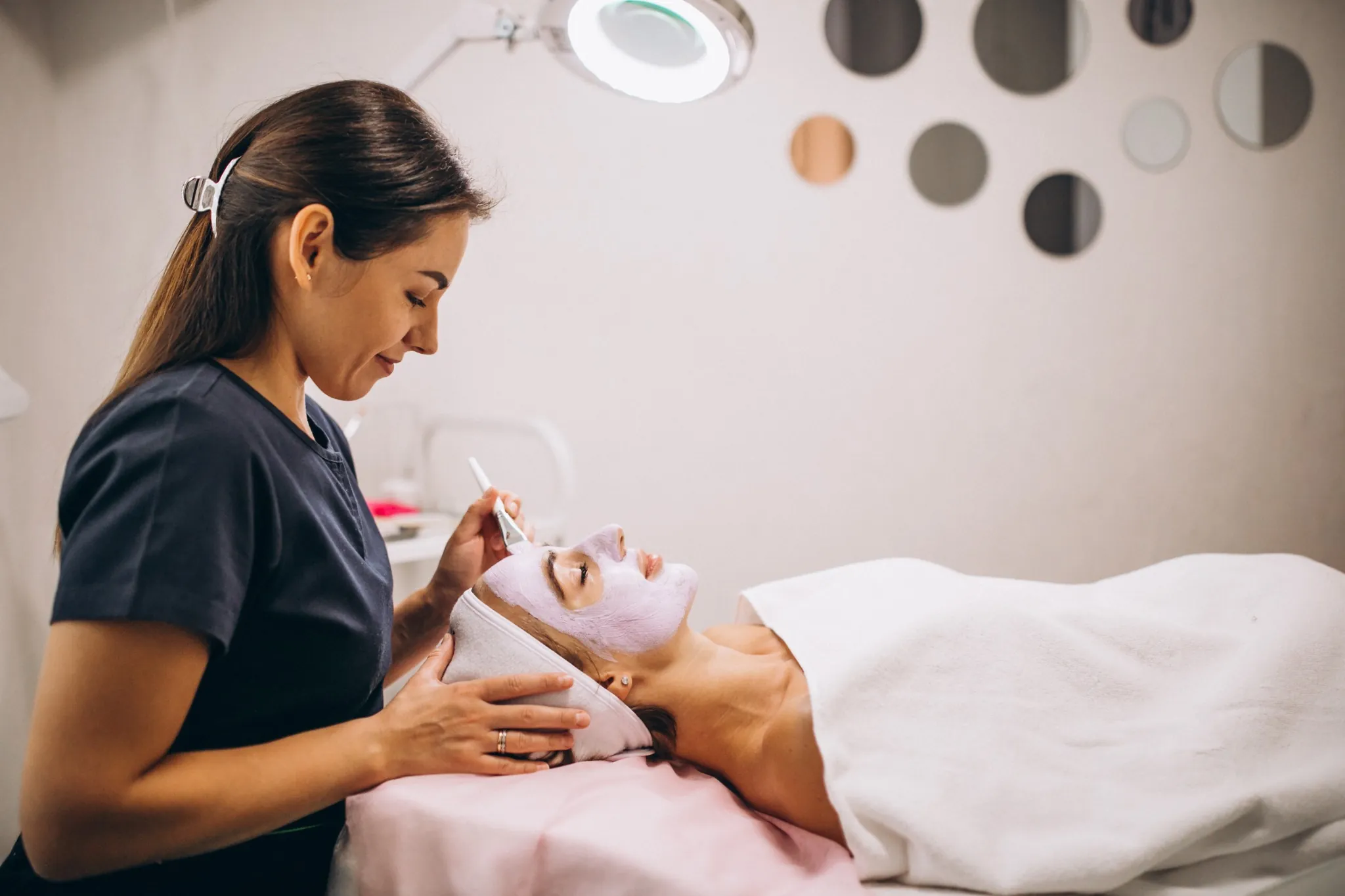 Profissional aplicando máscara facial em cliente relaxada em salão de beleza.