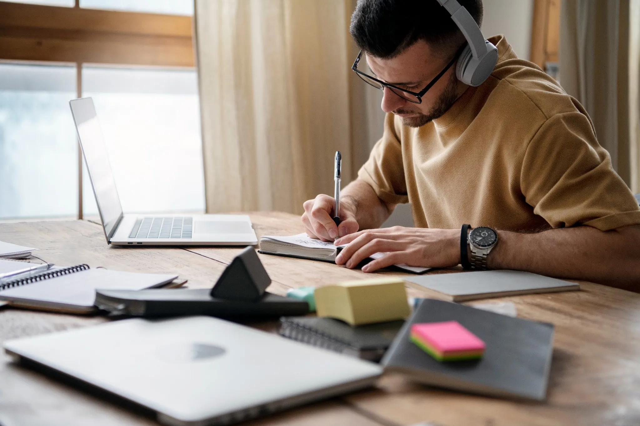 Jovem concentrado estudando em casa com caderno e laptop.