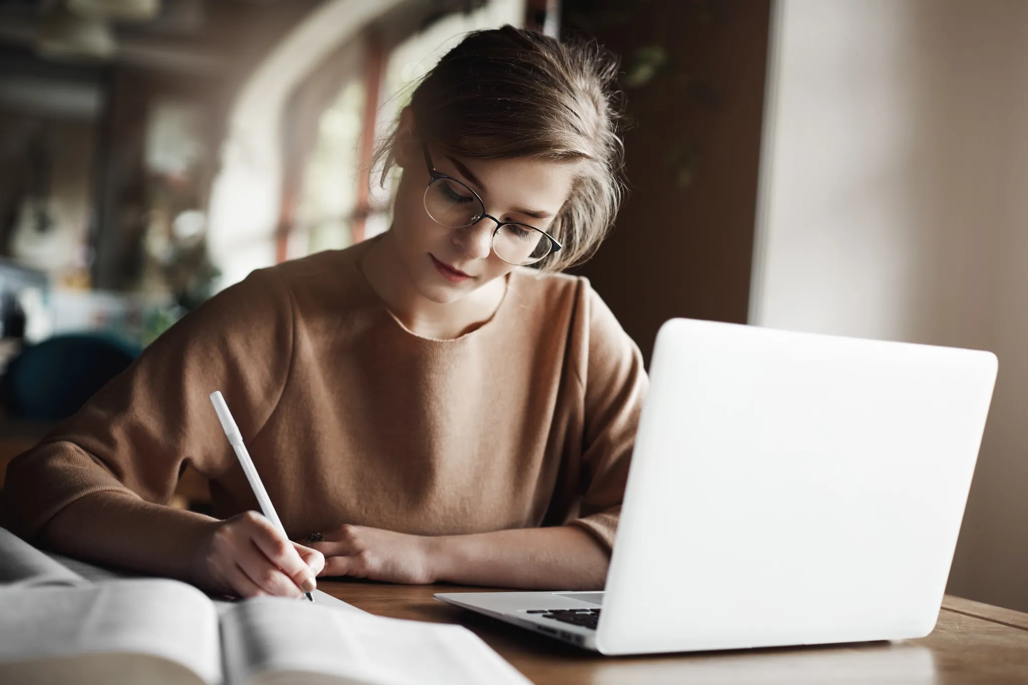 Jovem mulher estudando com laptop e caderno em ambiente aconchegante.
