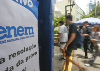 Estudantes andando em frente a um cartaz do ENEM em área urbana movimentada.