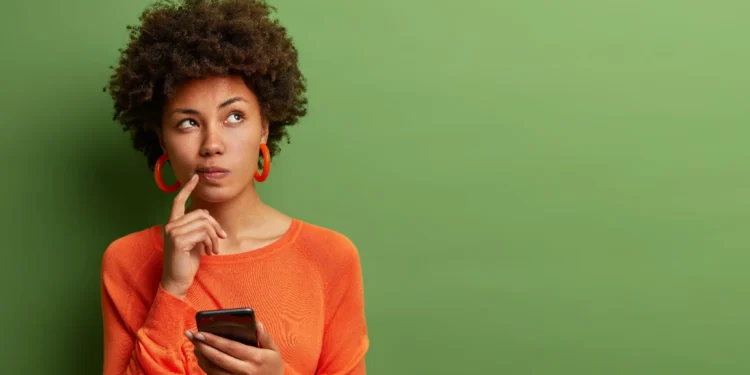 Mulher segurando celular com expressão pensativa em fundo verde.