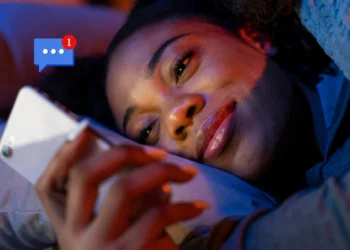 Mulher sorrindo enquanto lê uma mensagem no celular deitada na cama.