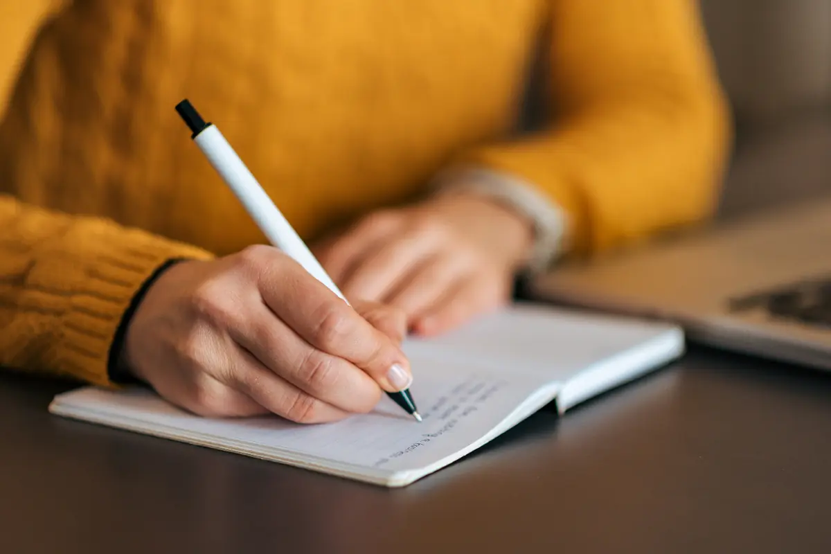 Pessoa escrevendo em caderno com caneta, vestindo blusa amarela
