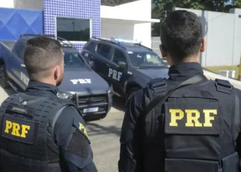 Dois policiais da PRF (Polícia Rodoviária Federal) observando seus veículos de patrulha.