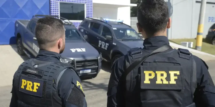 Dois policiais da PRF (Polícia Rodoviária Federal) observando seus veículos de patrulha.