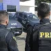Dois policiais da PRF (Polícia Rodoviária Federal) observando seus veículos de patrulha.