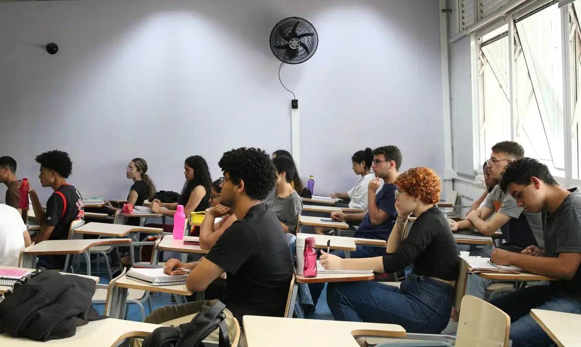 Estudantes atentos em sala de aula moderna, com cadernos e garrafas sobre as mesas.