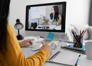 Pessoa estudando online com videoaula no computador e anotações em caderno