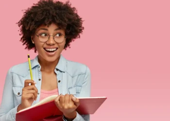 Estudante sorridente segurando caderno e lápis em fundo rosa.