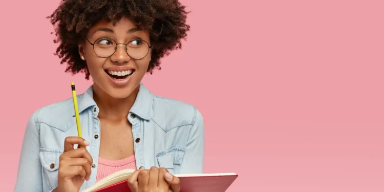 Estudante sorridente segurando caderno e lápis em fundo rosa.