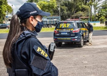Policial mulher rodoviário federal com arma ao lado de um veículo da PRF em estrada, aguardando ações de fiscalização.