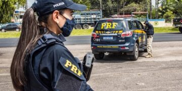 Policial mulher rodoviário federal com arma ao lado de um veículo da PRF em estrada, aguardando ações de fiscalização.