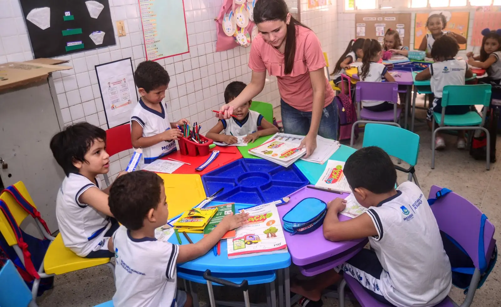 Professora auxilia alunos da educação infantil em sala de aula colorida.