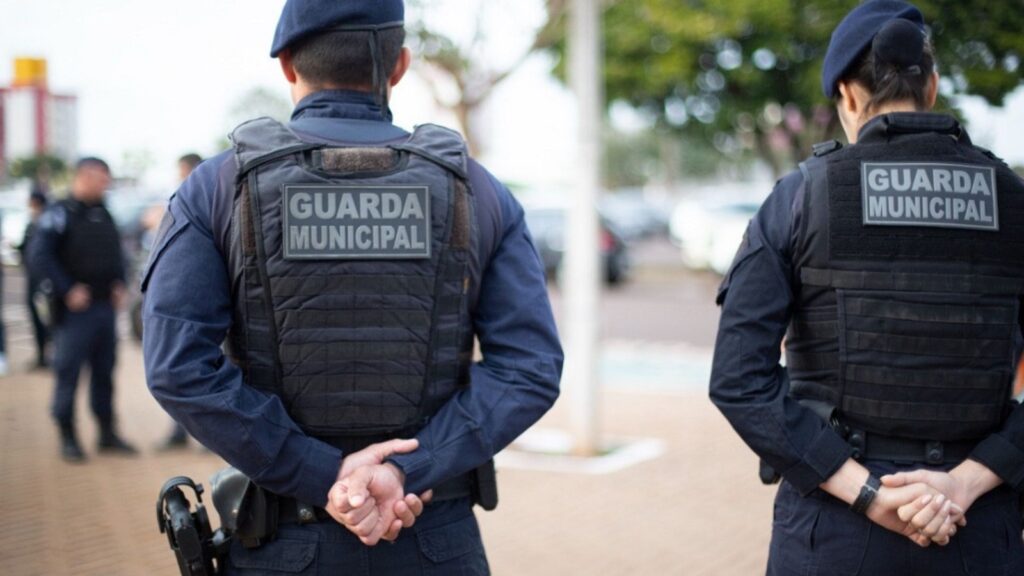Guardas municipais de costas, com coletes táticos e a identificação 'GUARDA MUNICIPAL', prontos para o serviço.