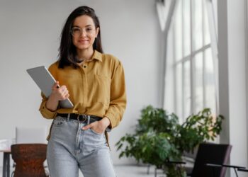 ovem profissional sorrindo e segurando um tablet em um escritório, representando as oportunidades de carreira.