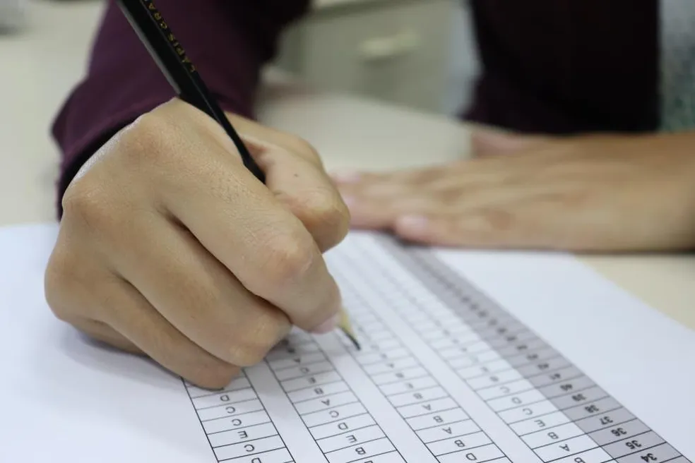 Mão preenchendo formulário com lápis, simbolizando preparação para concurso público.