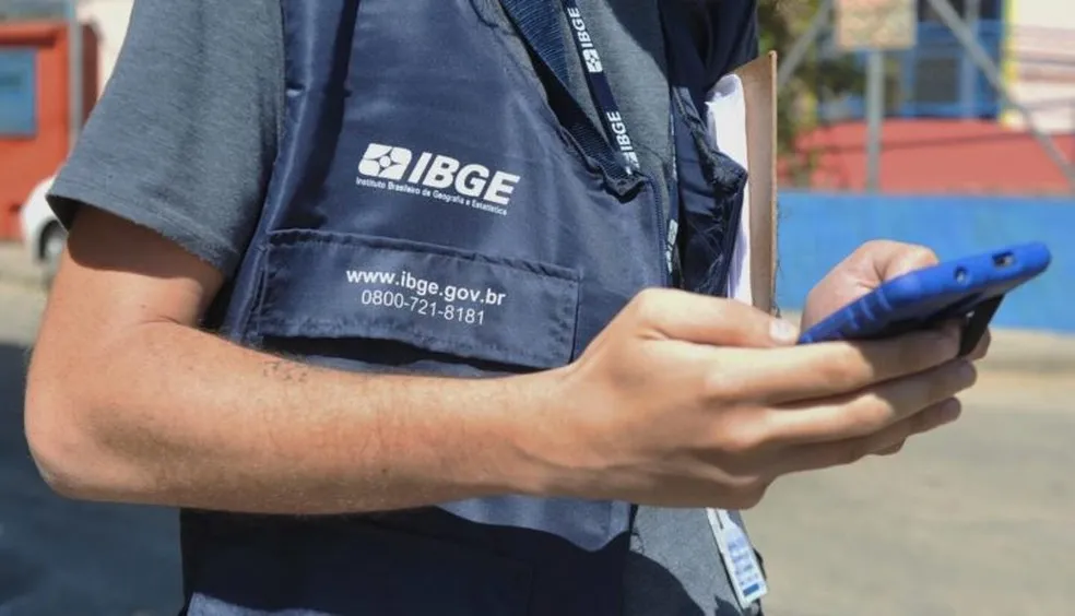 Funcionário do IBGE com colete e celular, realizando trabalho de coleta de dados para o censo.
