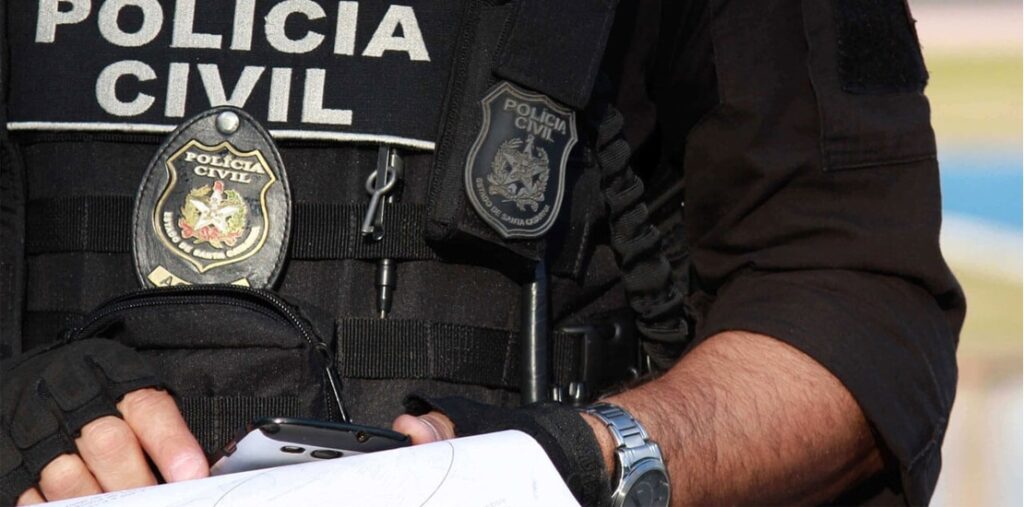 Polícia Civil MS em ação. Detalhe do uniforme da Polícia Civil de Mato Grosso do Sul com distintivo, colete e celular em mãos, simbolizando o trabalho investigativo.