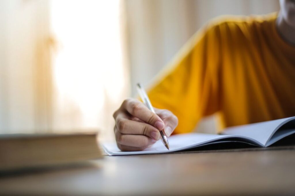 Pessoa em camiseta amarela escrevendo em um caderno com uma caneta, em uma mesa de estudos com um livro ao lado, representando os estudos para concursos. 
