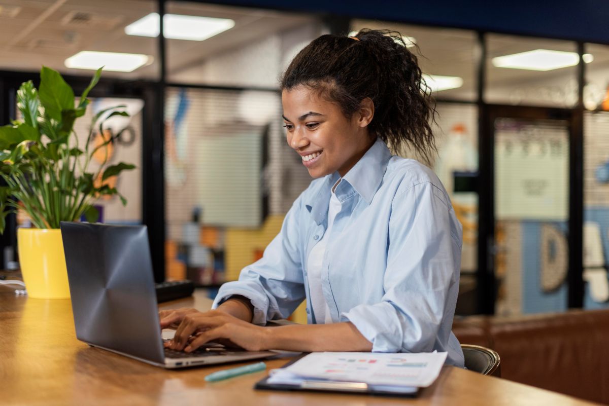 Jovem estagiária sorrindo e trabalhando em seu notebook em um escritório moderno.