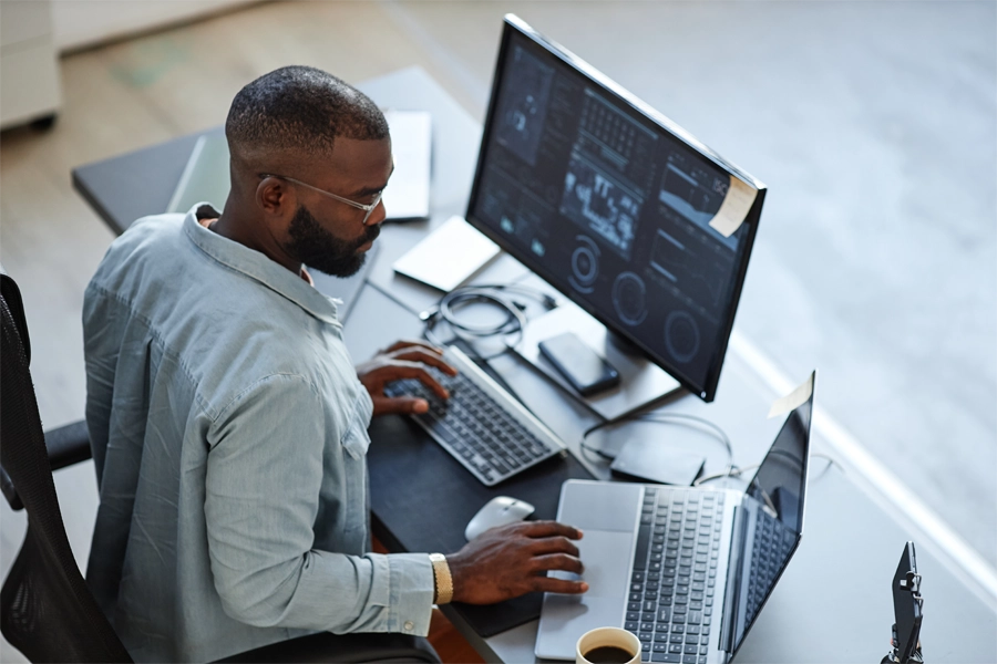 Profissional de TI trabalhando em dois computadores, com gráficos e dados exibidos na tela.