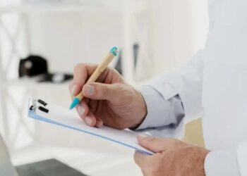 Mão segurando uma caneta e anotando em um bloco de notas, simbolizando inspeção médica ou exames de saúde.