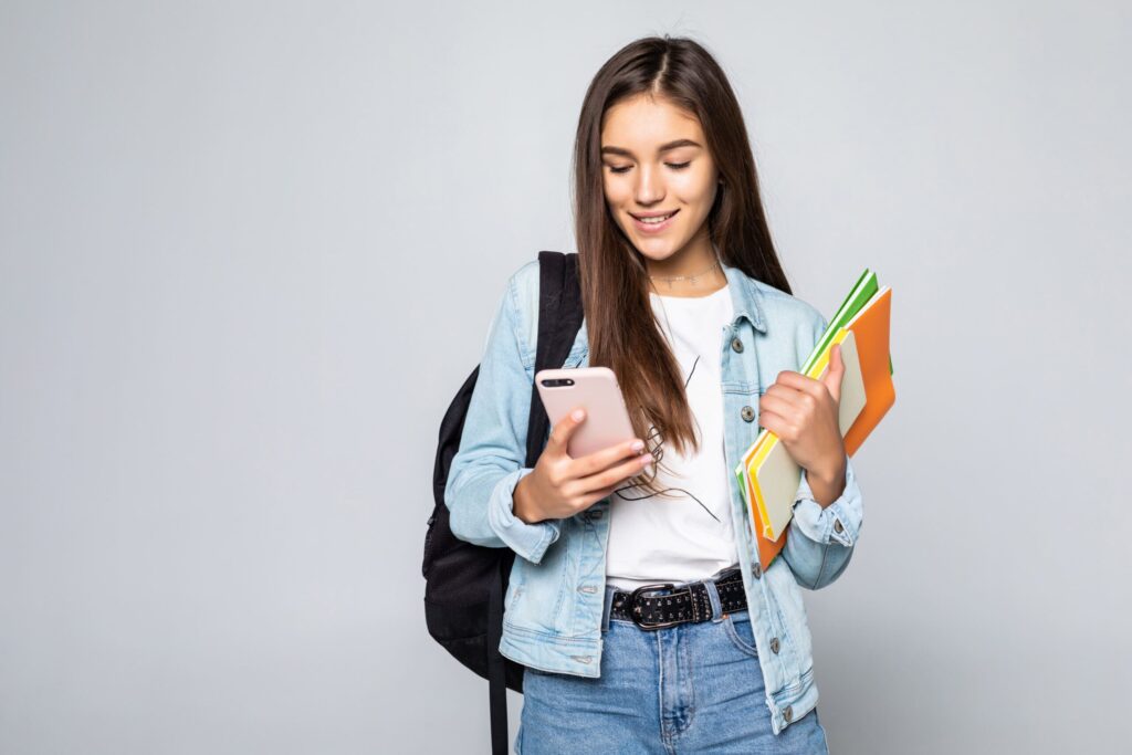 Jovem estudante sorrindo, com mochila, livros e celular na mão, simbolizando a busca por qualificação e o uso da tecnologia para o estudo.