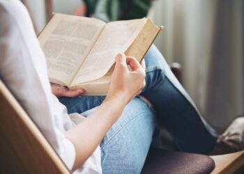 Pessoa lendo um livro com as mãos em uma poltrona confortável, representando a importância de leituras relaxantes e obras literárias alternativas para concurseiros recarregarem as energias e manterem a mente ativa durante a preparação.