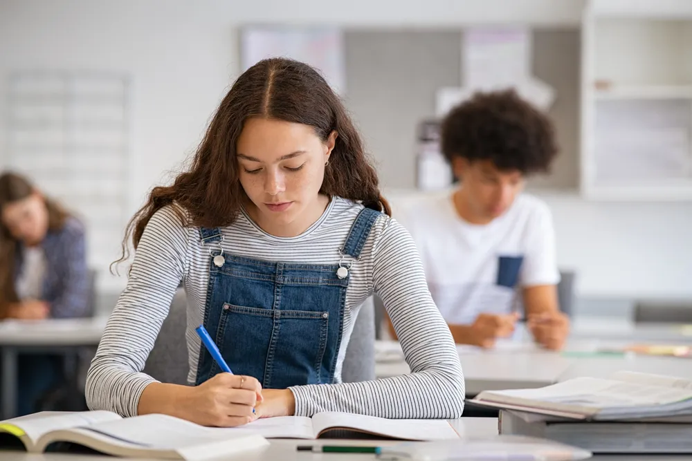 Jovem estudante com cabelo cacheado e macacão jeans escrevendo com caneta azul em um caderno ou apostila durante uma prova ou sessão de estudos intensa.