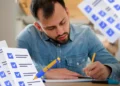 Homem sério usando camisa jeans escrevendo em caderno com papéis de checklist ao redor