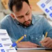 Homem sério usando camisa jeans escrevendo em caderno com papéis de checklist ao redor