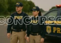 Dois policiais da Polícia Rodoviária Federal com uniformes completo em pé ao lado de viatura PRF