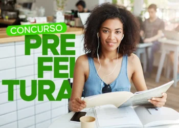 Mulher jovem estudando e sorrindo enquanto ouve música, com fones de ouvido, ao lado de uma mesa com materiais de concurso, com destaque para o concurso da Prefeitura de Joanópolis.