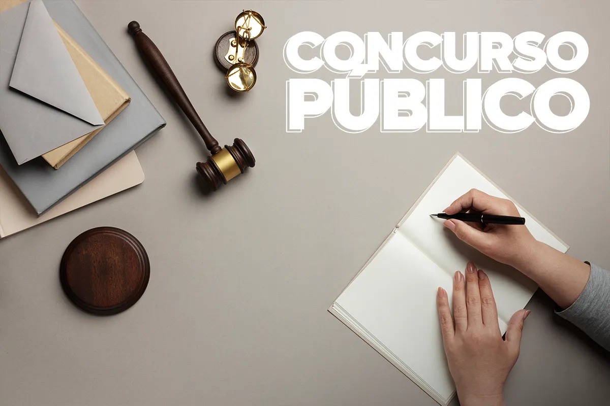 Mãos escrevendo em um caderno aberto, com um martelo de juiz e balança da justiça ao lado, representando a preparação para concursos públicos e processos seletivos no setor paulista