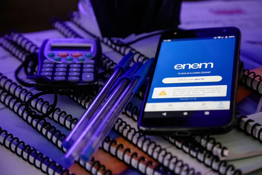 Celular sobre cadernos e canetas com a tela de login "Enem - Entrar com gov.br", representando o acesso online ao Cartão de Confirmação do exame.