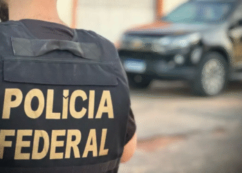 Agente da Polícia Federal com colete e viatura ao fundo.