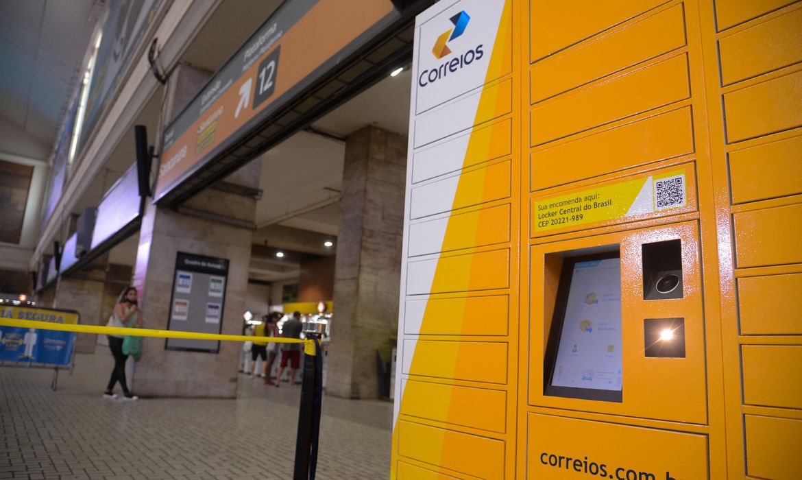 Locker dos Correios amarelo para retirada de encomendas em estação com pessoas ao fundo