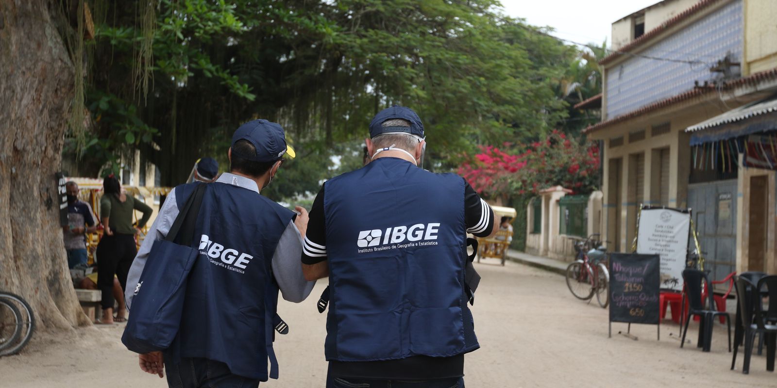  Dois recenseadores do IBGE vestidos com coletes e bolsas azuis caminhando em área residencial