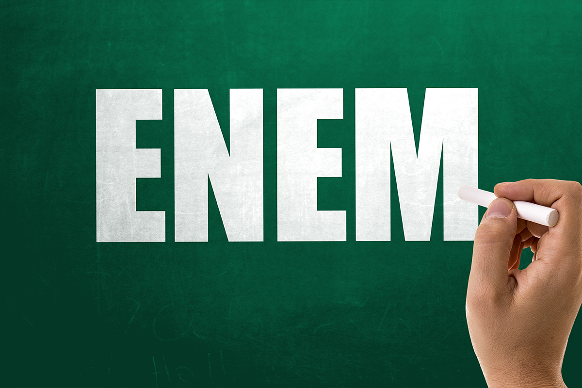 Mão escrevendo a palavra ENEM em letras grandes brancas sobre quadro verde com giz branco
