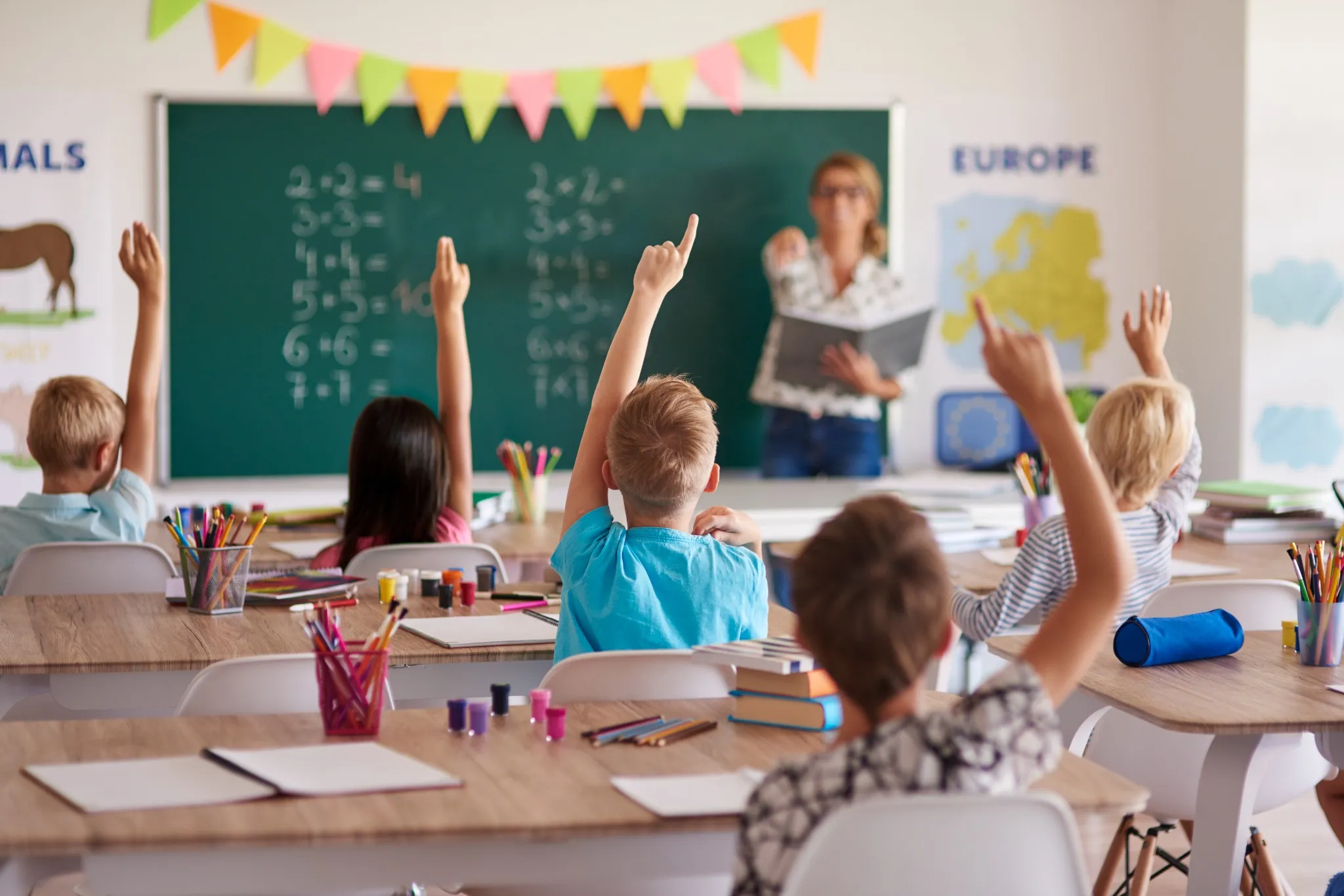 Alunos levantando a mão em sala de aula enquanto a professora ensina matemática, ilustrando a aplicação de padrões mínimos de qualidade para a educação básica